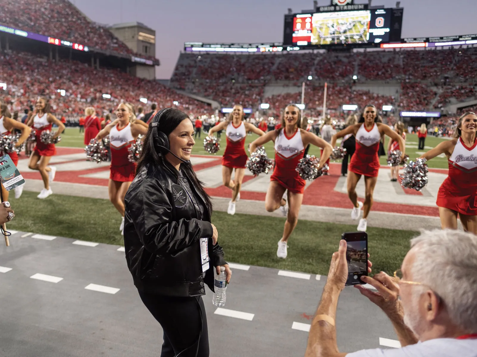 Melissa McGhee, a short white woman with long dark hair, smiles as she coaches the dance and cheer teams on the sidelines of an Ohio State football game. In Ohio Stadium, she wears a leather Ohio State coat and a headset.