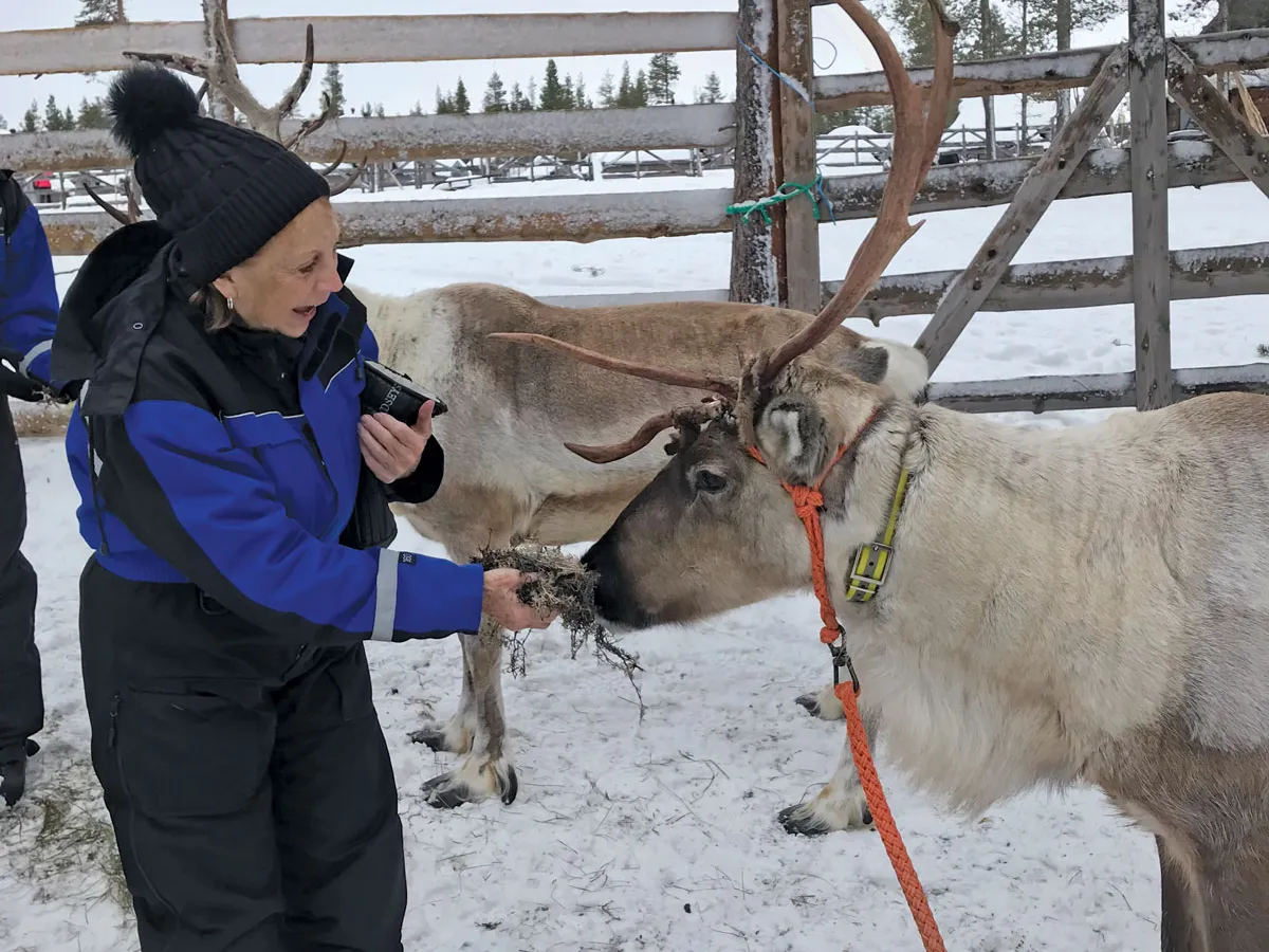 A white woman in snow gear feeds a reindeer on a trip. They&#039;re surrounded by snow.