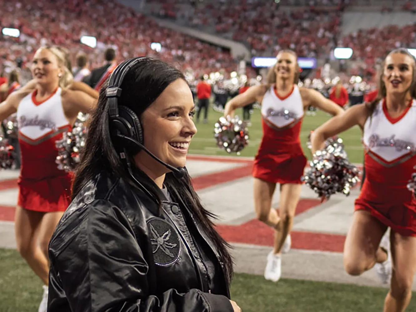 Melissa McGhee, a short white woman with long dark hair, smiles as she coaches the dance and cheer teams on the sidelines of an Ohio State football game. In Ohio Stadium, she wears a leather Ohio State coat and a headset.