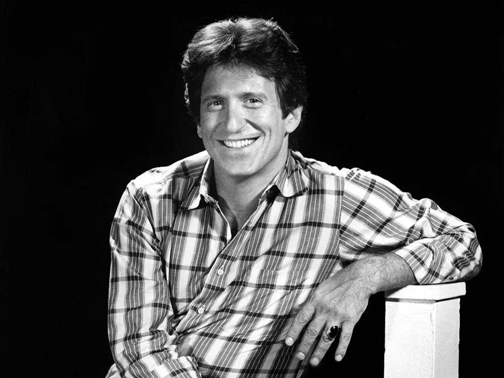 Michael Ochs smiles for a portrait as a younger man. A white man, he has thick hair, a big smile and wears a plaid shirt and jeans.