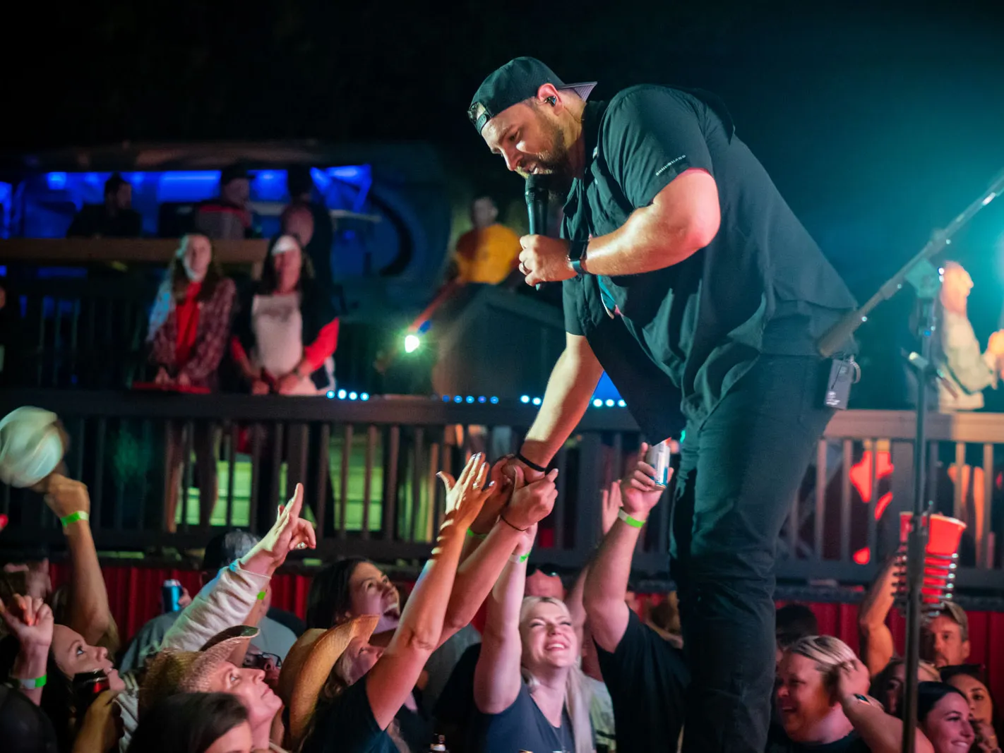 A man holding a mic on stage reaches down to clasp hands with adoring audience members. He wears all black and a backwards baseball cap.