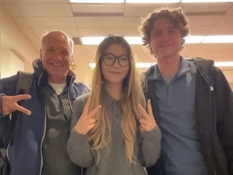 Three people grin as they pose for a casual, selfie-style photo. A grandpa and high school student stand with their TA between them.