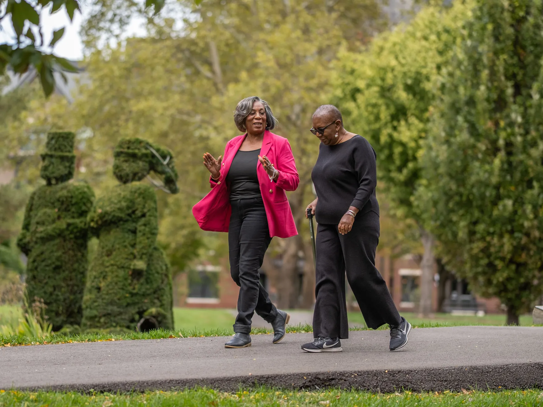 Two black ladies smile as they chat and walk through a famous park in downtown Columbus. One uses a cane and wears head-to-toe black. The other wears a bright pink suit jacket over her black outfit.