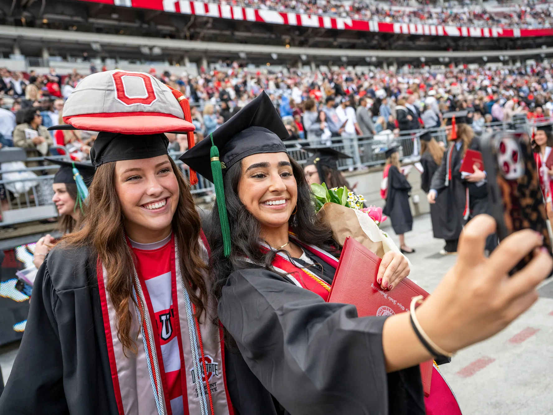 Two people in graduation caps and gowns take a selfie on a football stadium field, with one holding a diploma and bouquet and the other wearing Brutus’ giant cap on top of her graduation cap. A crowd of graduates and guests fills the stands in the background.