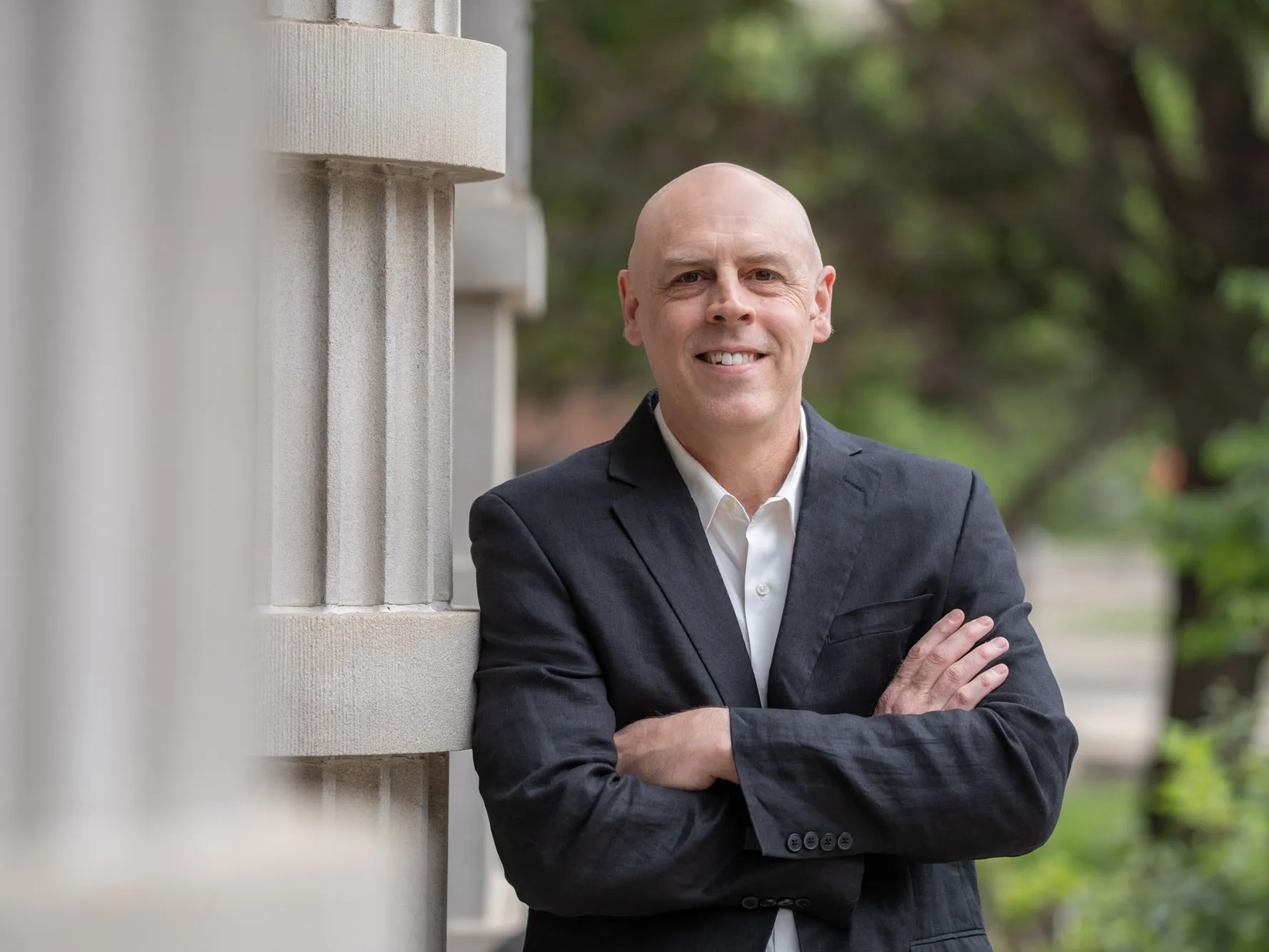 A professor smiles as he stands outdoors leaning against a column with arms crossed, wearing a collared shirt and suit jacket. He is a white man with a shaved head. Trees and a walkway appear in the background of the photo.
