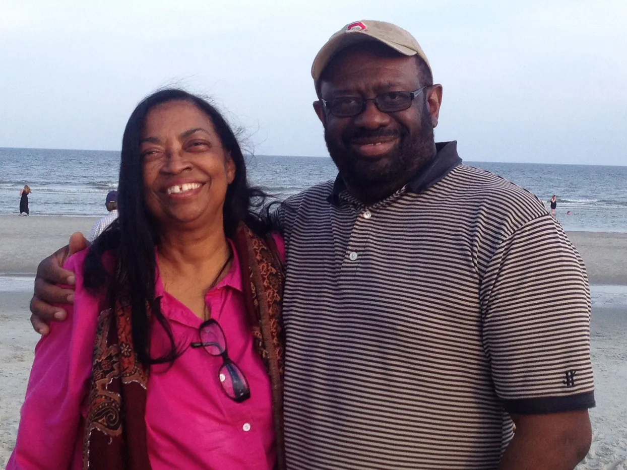 A husband and wife smile as they stand close together on a sandy beach with the ocean and a few distant beachgoers in the background.
