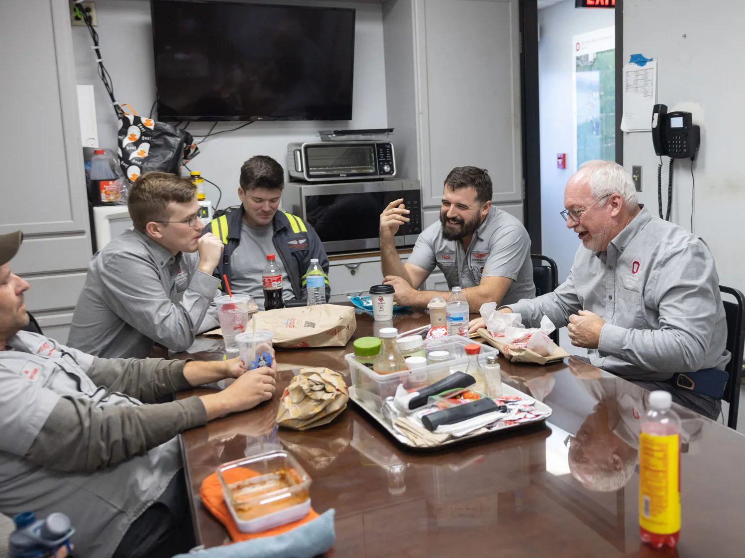 At least five men people sit around a round table in a break room, eating lunch. The table has fast‑food bags, sandwiches, and other meal items on it. The individuals are wearing matching work uniforms and are seated in a small room with cabinets, a wall clock, a microwave, and a television visible in the background.