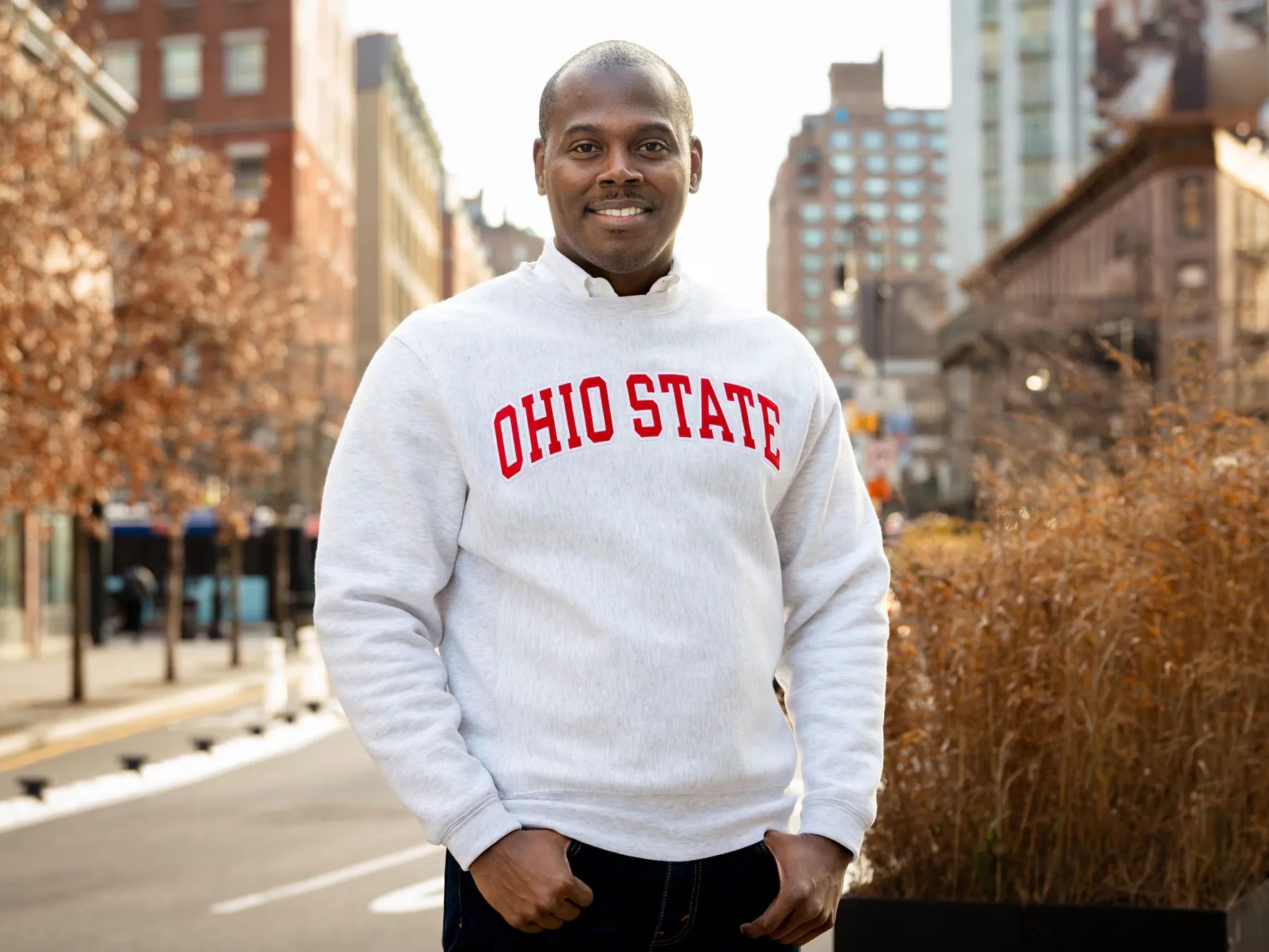 An African-American man smiles as he stands on a city street wearing a sweatshirt with the text “Ohio State” across the front, with thumbs tucked in his pockets and buildings and trees in the background. He looks like he's interested in what you have to say.