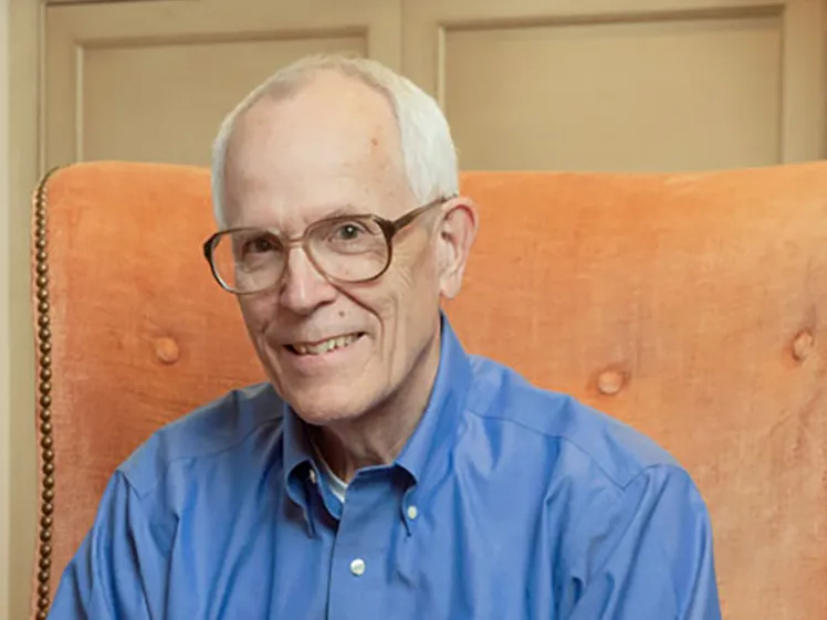 Andy Ross smiles as he sits in a sophisticated upholstered armchair with hands loosely folded. He wears a button down, dress pants and glasses, and he looks like someone who would be interesting to have a conversation with.