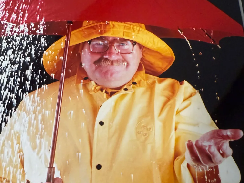 A person wearing a yellow raincoat and holding a red umbrella stands in falling water, with one hand extended outward to catch the rain. He is John Switzer and he has a mischievous smile, big glasses and a big mustache that hint at his humor