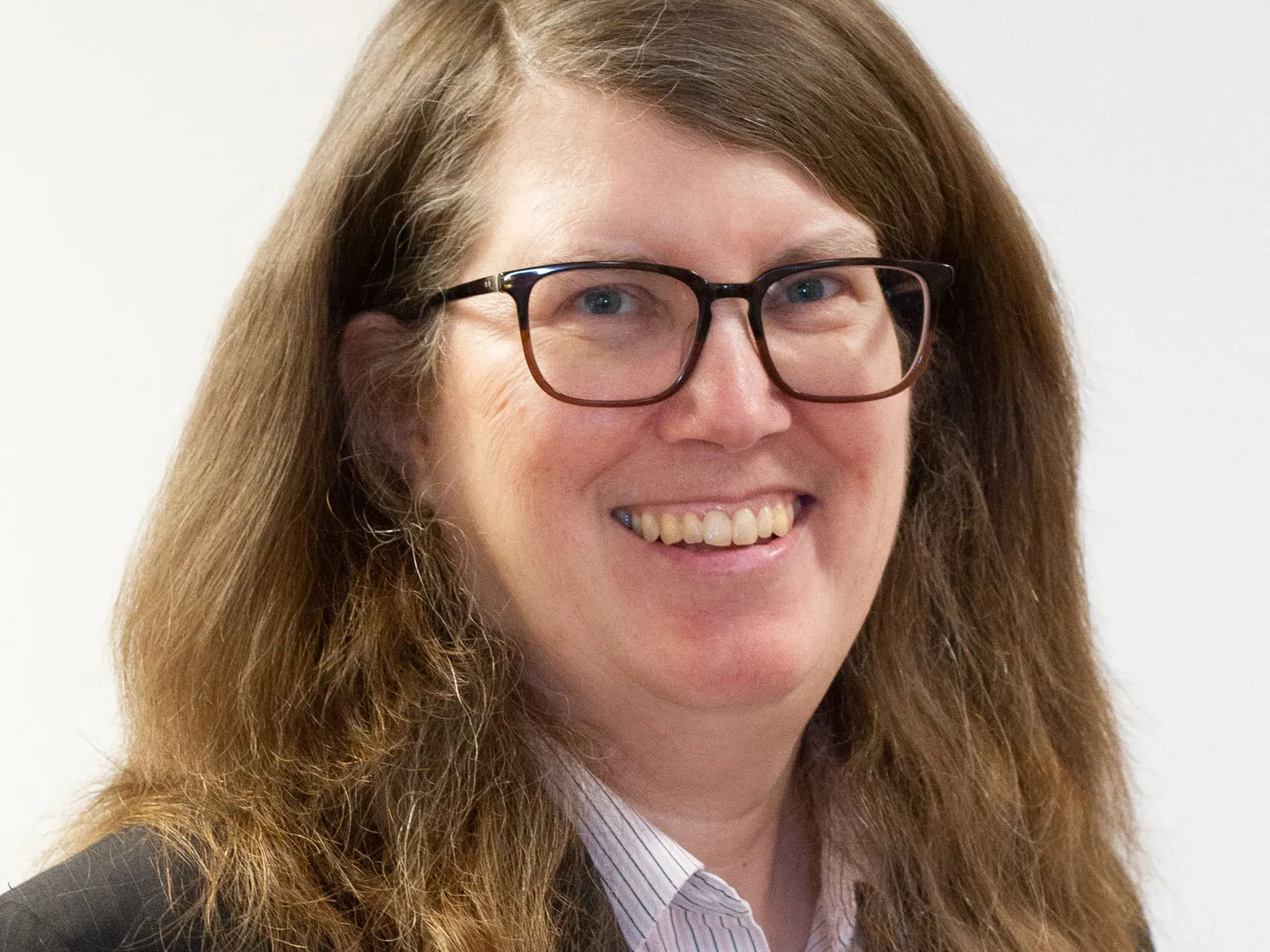 With long wavy hair and wearing glasses, a dark blazer and a beaded necklace, Ann Christy grins in a genuine way in this headshot.
