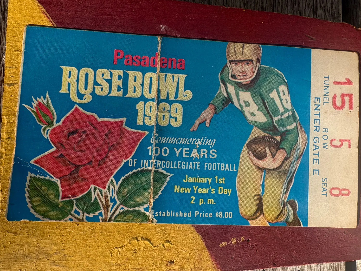 A 1969 rose bowl ticket sits on a slab of wood painted in maroon and yellow.