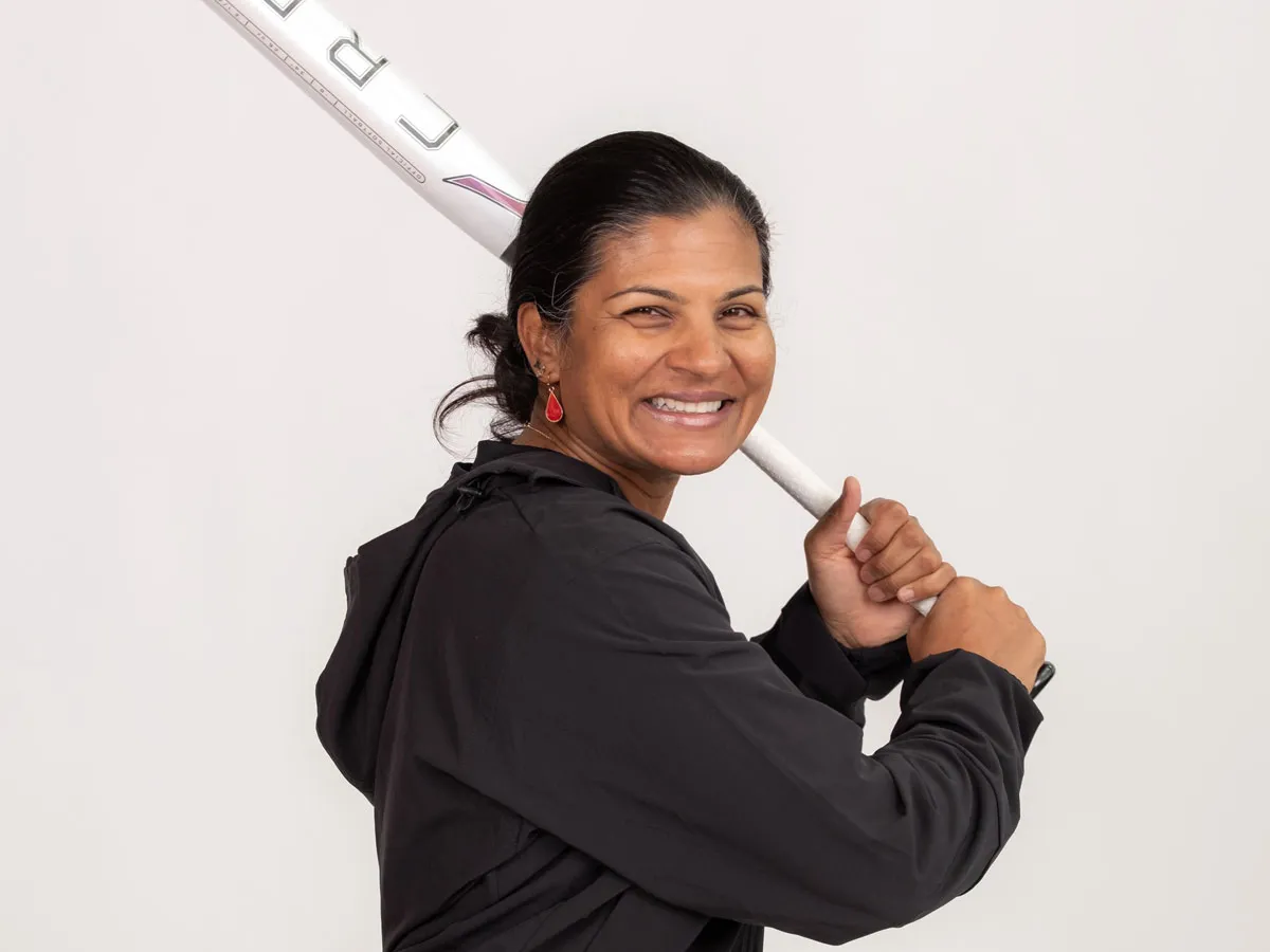 Kirin Kumar giggles as she poses in a batting stance, holding a baseball bat over one shoulder as if she’s getting ready for a pitch. 