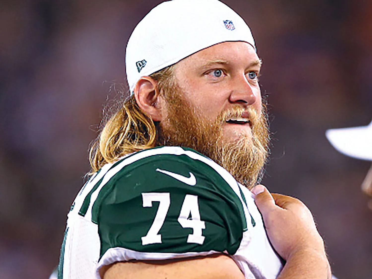 A football player with long blond hair stands along the field in a white and green uniform with the number 74, holding one arm across his chest pads. He is Nick Mangold and he looks pleased by what he’s watching.