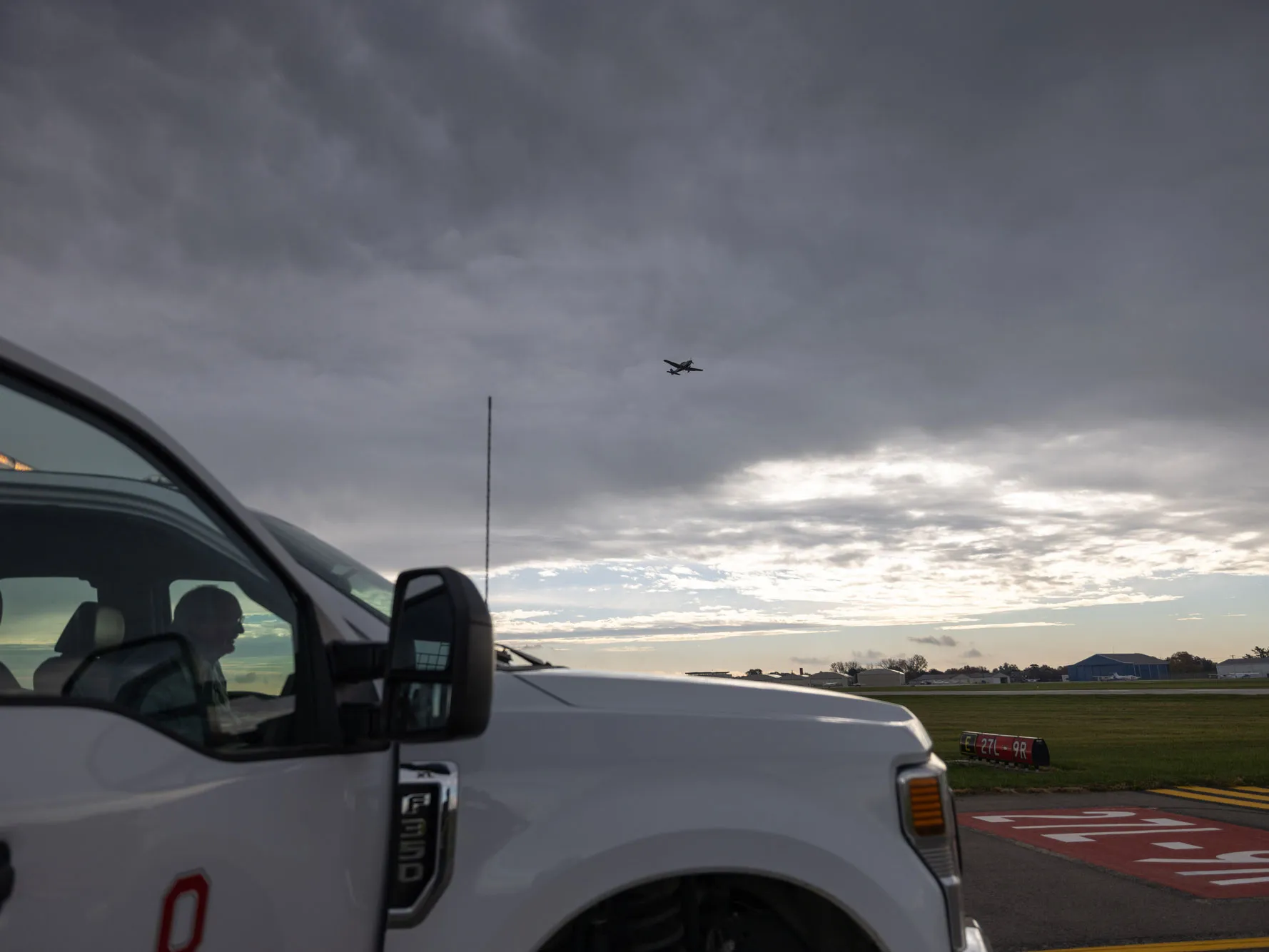 A person sits inside a parked truck near an airport runway, with part of the person's face visible through the window. A plane flies overhead beneath a cloudy sky.