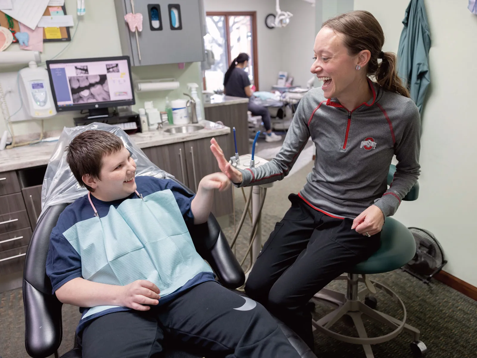 A dentist gives a high five to a young patient's fist bump. He sits in a chair with the paper bib people wear to be examined. And she wears an Ohio State shirt. They're both grinning as the watch each other.