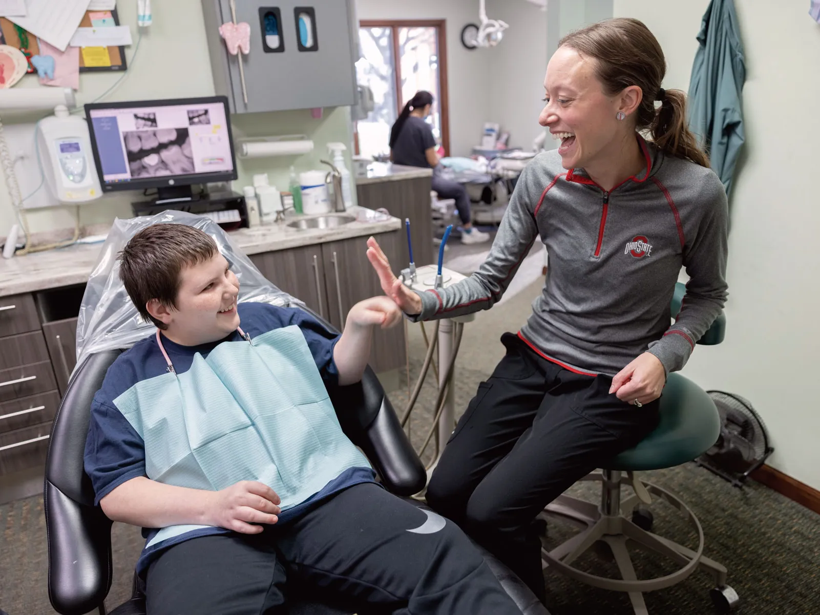 A dentist gives a high five to a young patient's fist bump. He sits in a chair with the paper bib people wear to be examined. And she wears an Ohio State shirt. They're both grinning as the watch each other.