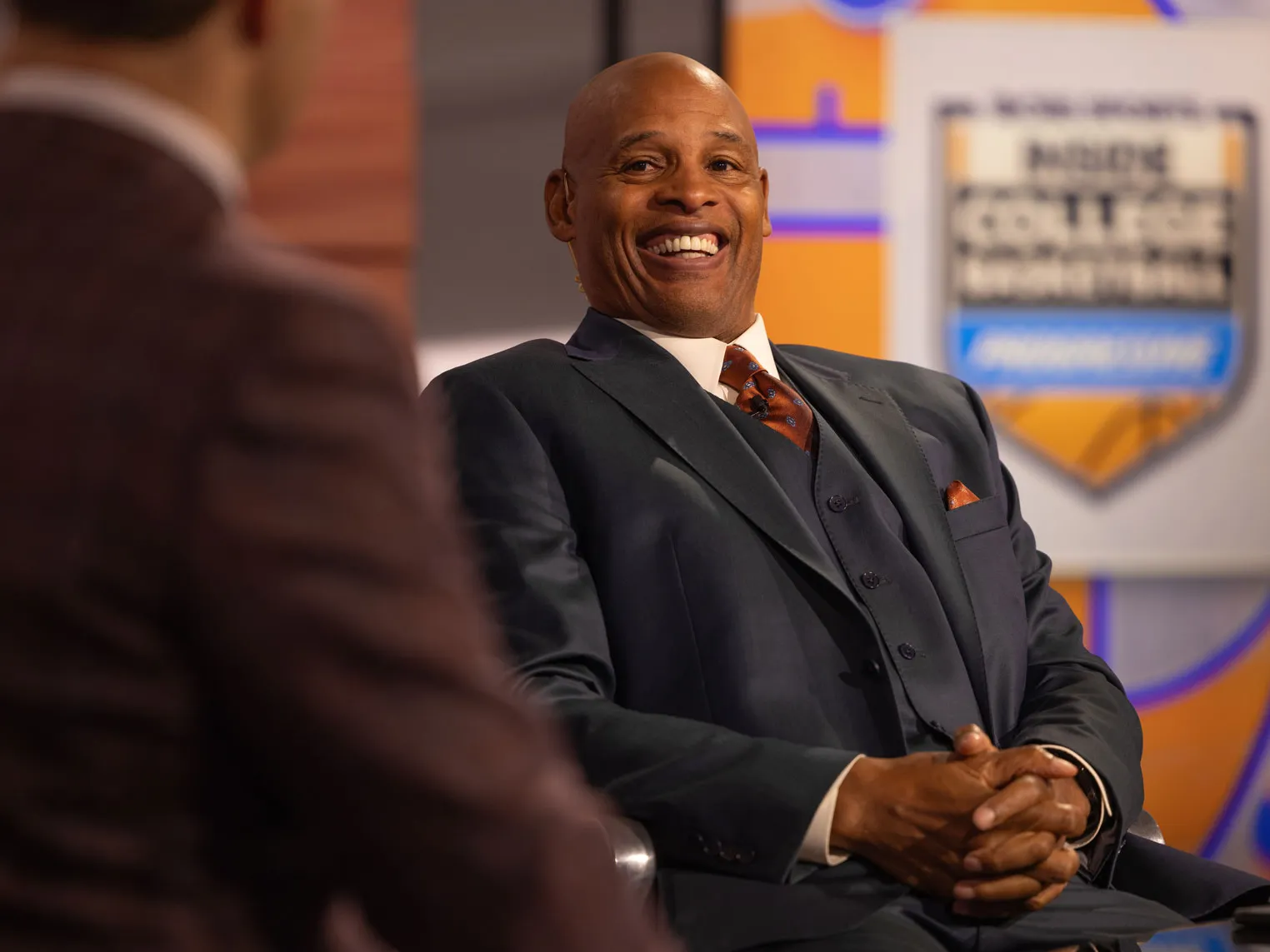 The besuited Clark Kellogg grins during an on camera interview in a studio, with another person visible in the foreground and a sports themed backdrop behind them.