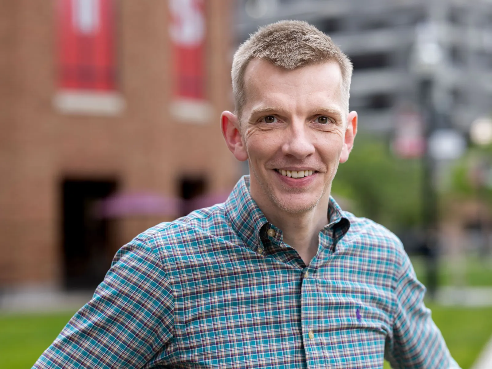 Angus Fletcher, a white man with neatly trimmed short hair, smiles and wears a patterned button-up shirt while standing outdoors with hands on hips. A brick building and a campus background with greenery appear behind him.