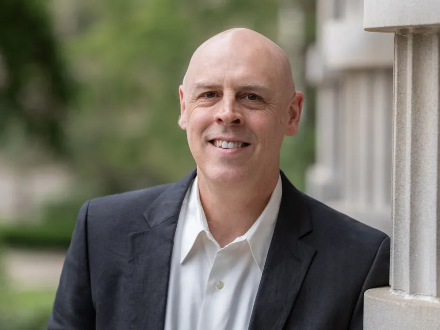 A professor smiles as he stands outdoors leaning against a column with arms crossed, wearing a collared shirt and suit jacket. He is a white man with a shaved head. Trees and a walkway appear in the background of the photo.