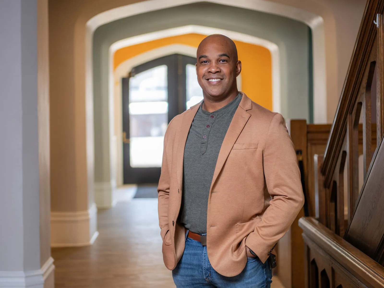 A Black man smiles as he stands in a hallway with hands in pockets, wearing a sport jacket and jeans, positioned near a wooden staircase with an arched doorway visible in the background. 