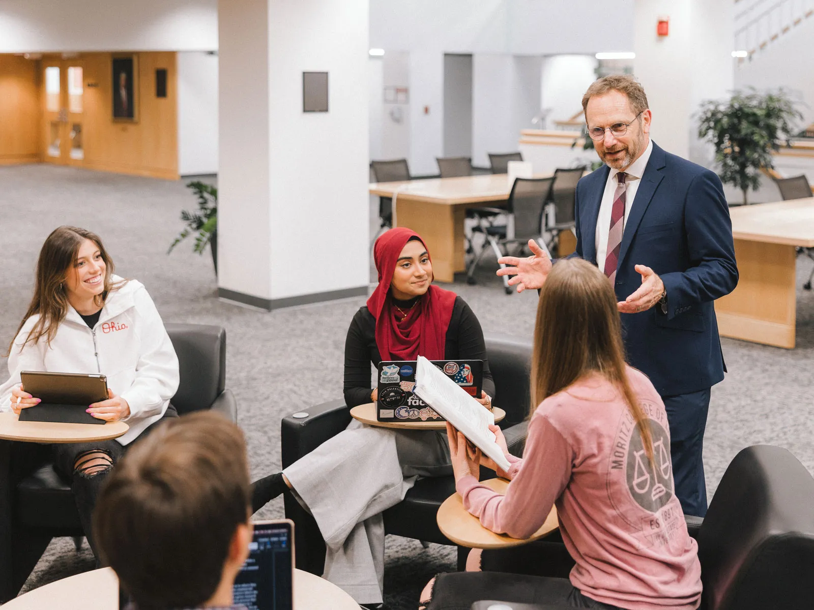 A group of law students sit together in a lounge-style area with small tables, using laptops and tablets, while Professor Dennis Hirsch stands and speaks with them. He’s talking with his hands as he explains a point.