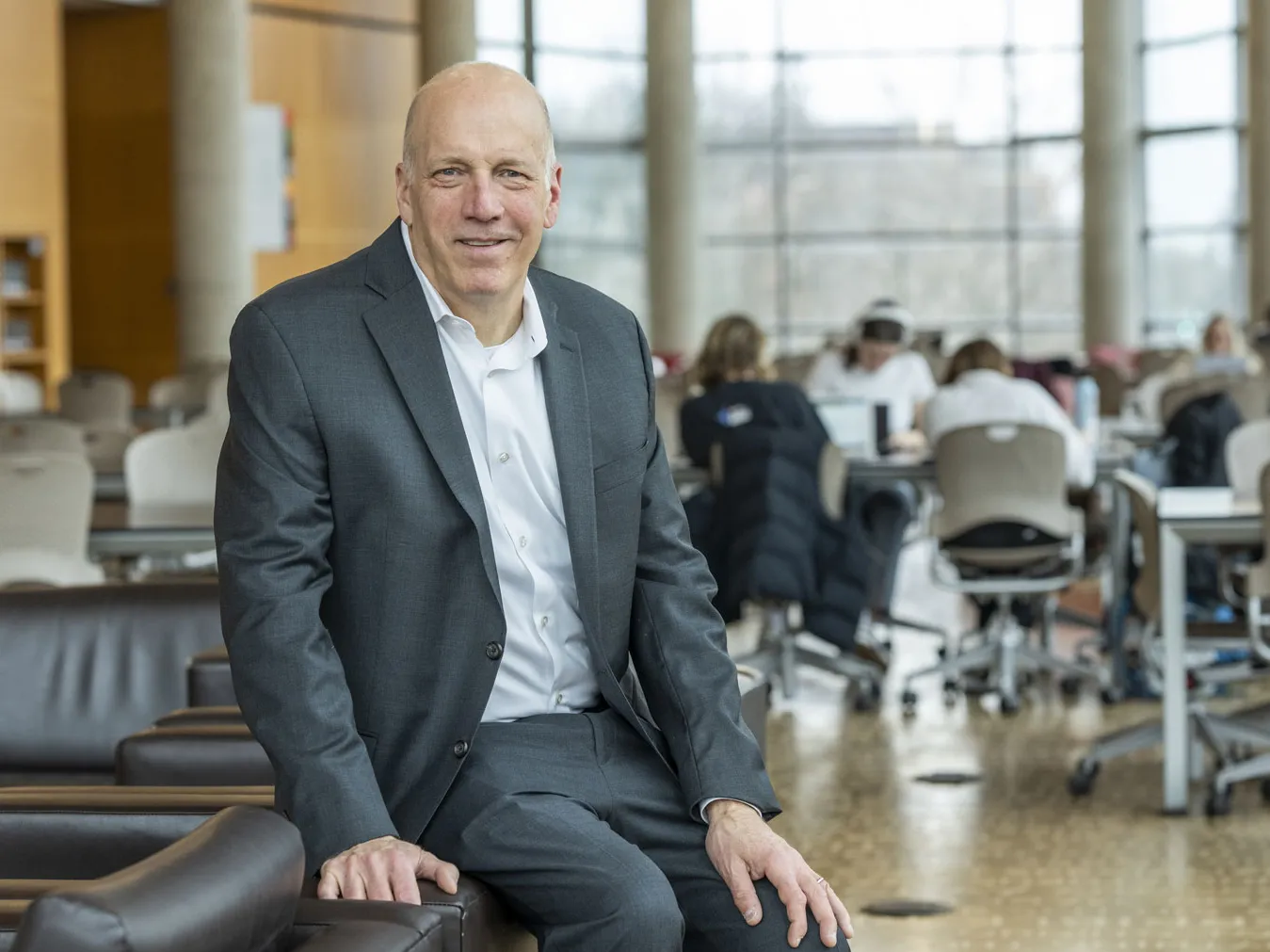 An older white man wearing a suit smiles as he sits on the edge of an upholstered chair in a spacious study room inside Thompson Library. Groups of people work at tables in the background and a full wall of windows and columns curves around the far side of the room.