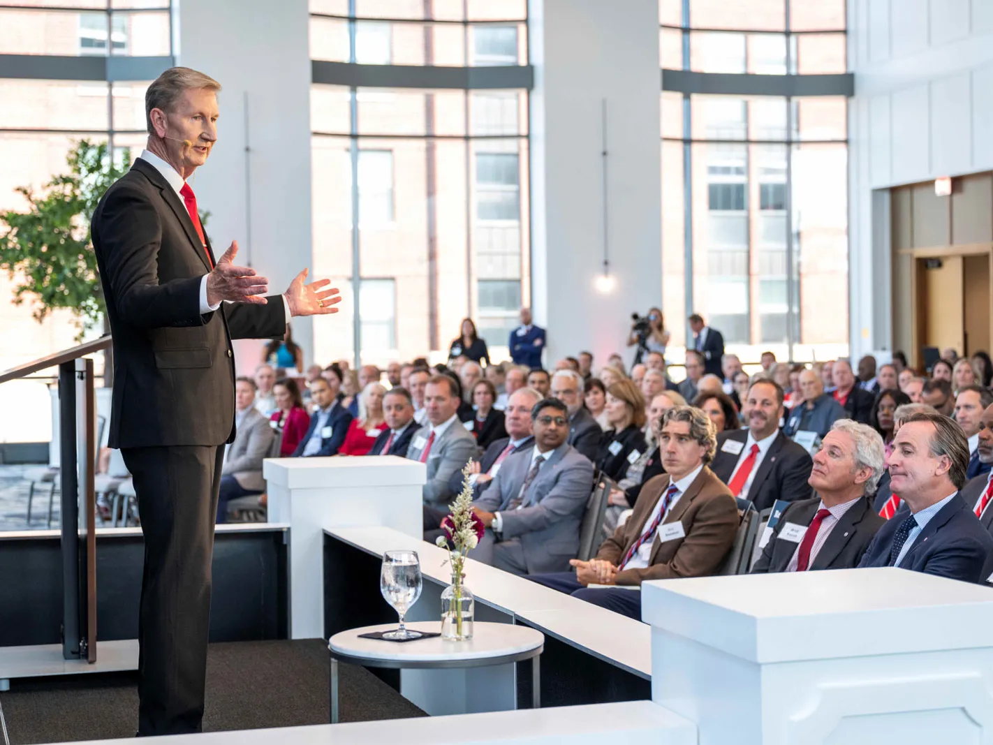 Ohio State President Ted Carter gestures as he speaks to an audience in a bright event space with tall windows. Attendees sit in rows facing him, listening with rapt attention. They’re all wearing suits or similar formal attire. 