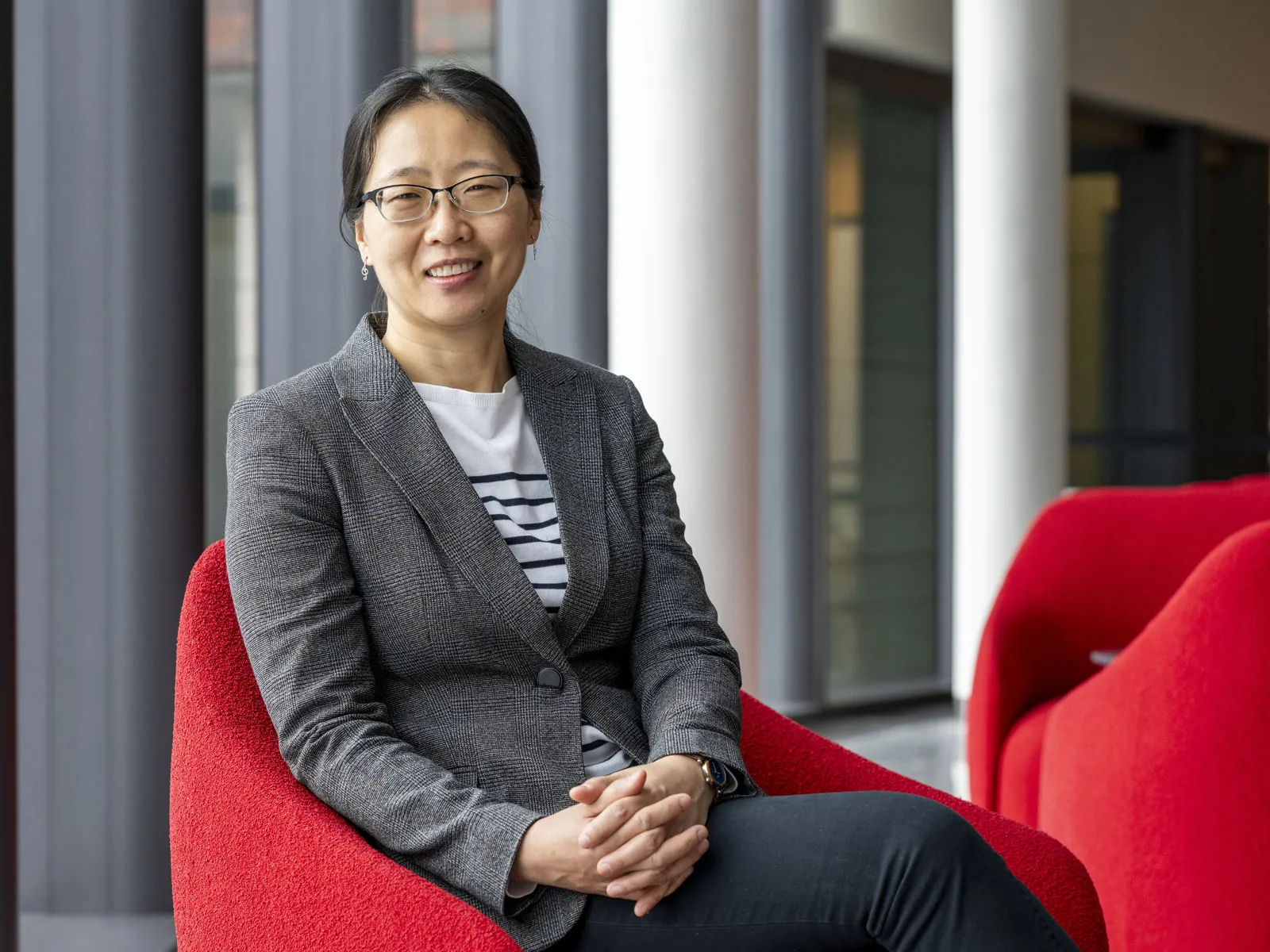 An Asian woman smiles as she sits in a modern indoor space on a curved chair, wearing a fitted blazer over a striped shirt with hands resting together. Tall windows, stately columns and additional chairs are visible in the background.