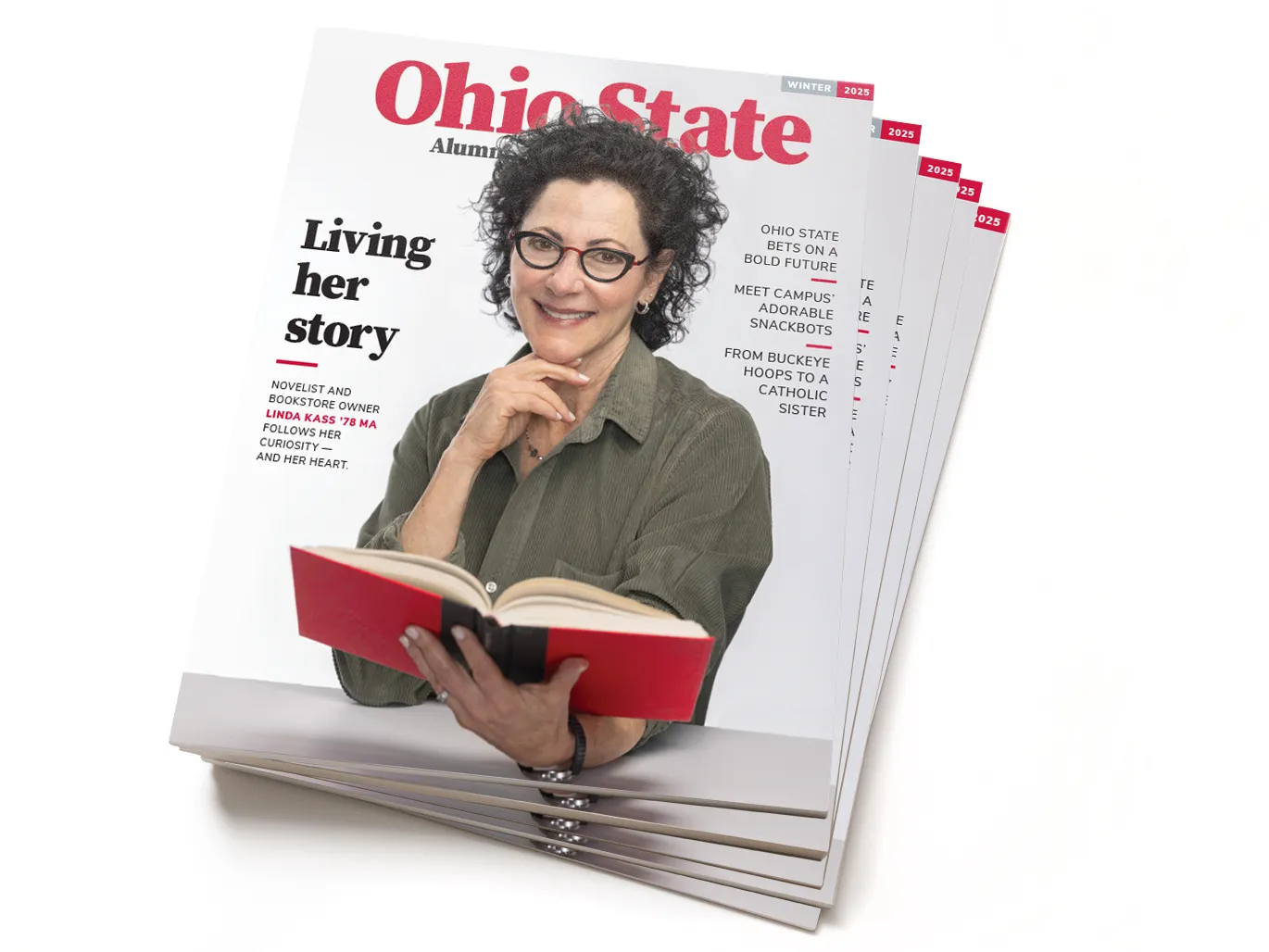 This is a stack of the Winter 2025 edition of Ohio State Alumni Magazine. The cover shows alumnus Linda Kass holding a book. Her other hand is tucked just beneath her chin. She’s a white woman with curly dark hair and glasses, and she looks happy, smart and well put-together. Her story is titled “Living her story” and other text on the cover says “Ohio State bets on a bold future”; “Meet campus’ adorable snackbots”; and “ From Buckeye hoops to a catholic sister”