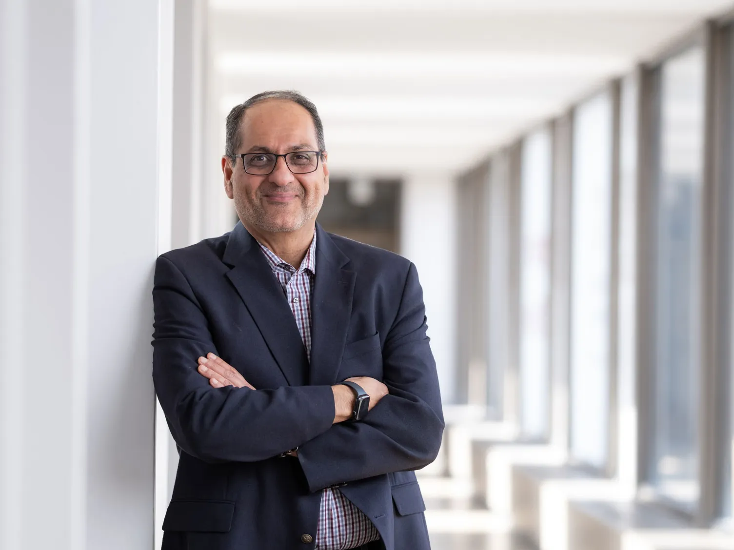Ness Shroff, a man of Indian descent, smiles as he leans against a wall in a bright hallway at Ohio State. He has arms crossed and wears glasses, a suit jacket and patterned shirt. He looks like someone who would be fascinating to have a discussion with. Large windows line the corridor in the background.