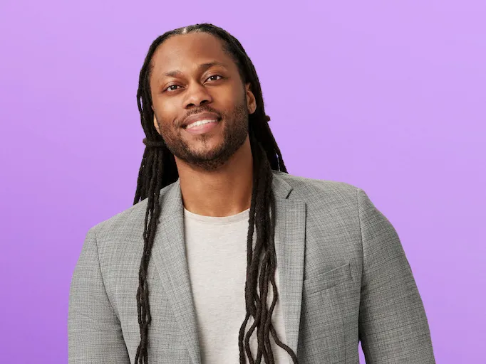Victor St. John, a black man with long dreadlock hair, wearing a blazer and jeans, looks friendly and studious—like he'd be the coolest professor—as he poses for this photo.  