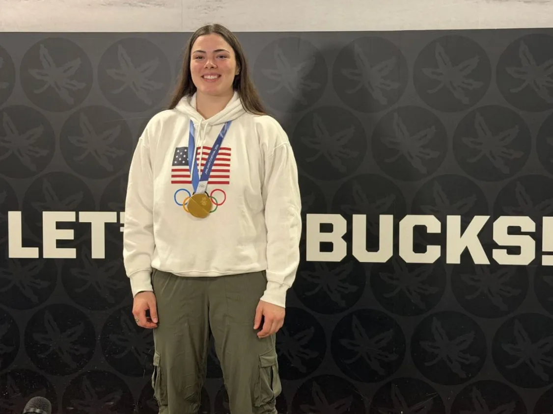 The women' hockey player Joy Dunne smiles as she wears a gold medal and stands in front of a wall that says "let's go Bucks!"