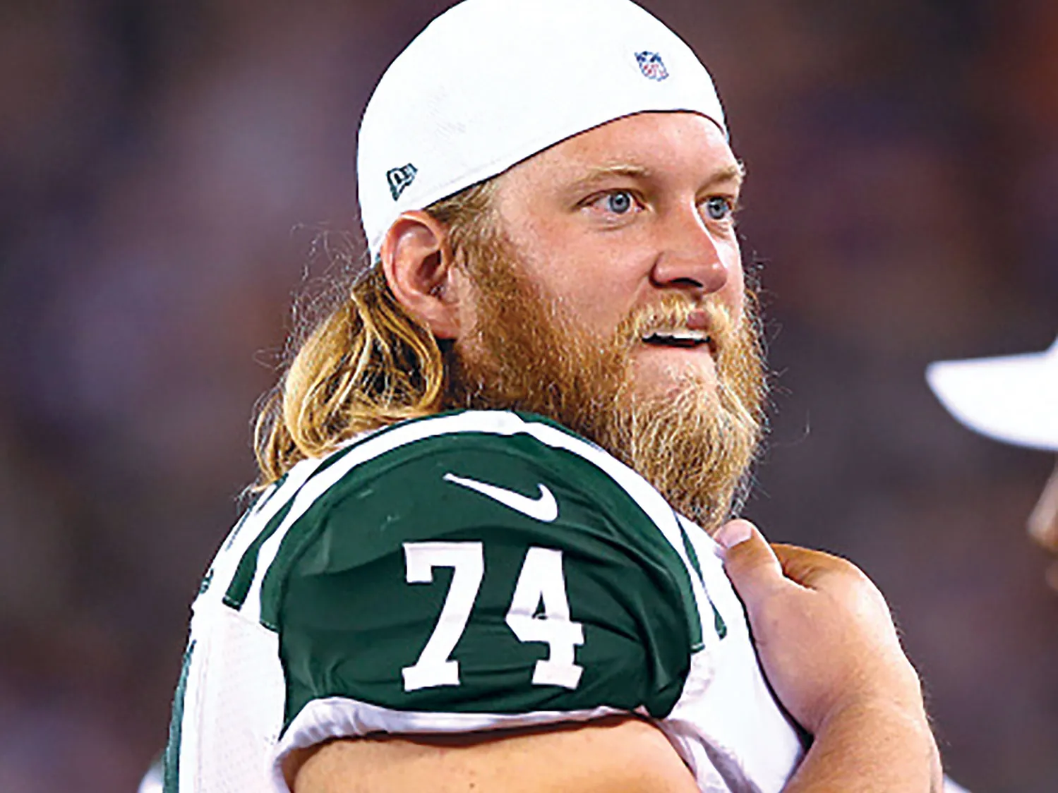 A football player with long blond hair stands along the field in a white and green uniform with the number 74, holding one arm across his chest pads. He is Nick Mangold and he looks pleased by what he’s watching.