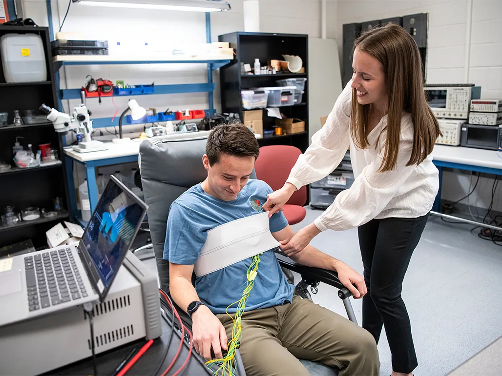 A young woman with l ong hair smiles as she hooks up a young man to a monitoring system that wraps a wide band around his chest. A computer and shelves of components are nearby.