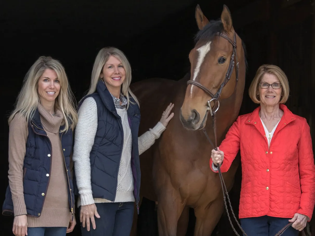 Three people stand beside a horse inside a barn entrance, with the mom holding the horse’s lead rope and her two grown daughters standing on the other side of the horse. They all smile as they pose.