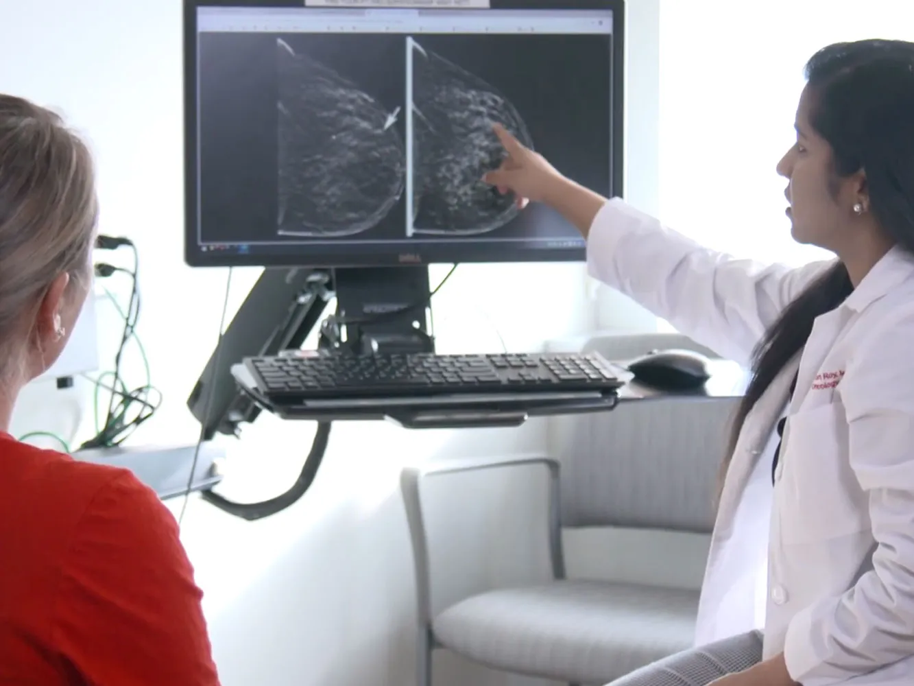 A medical professional in a white coat points to breast imaging scans on a computer monitor while another person sits nearby, looking at the screen.