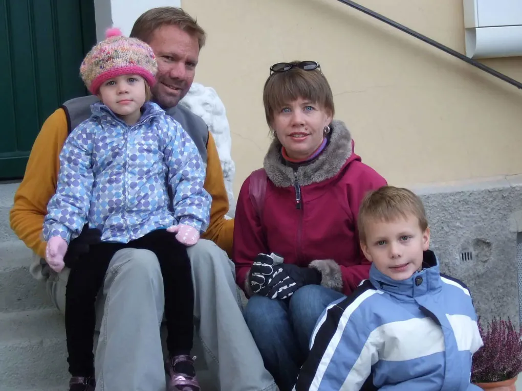 A father sits on stairs with his 3-year-old daughter on his lap, his wife by his side and his son a couple of steps down. They're all smiling as the pose for the picture.