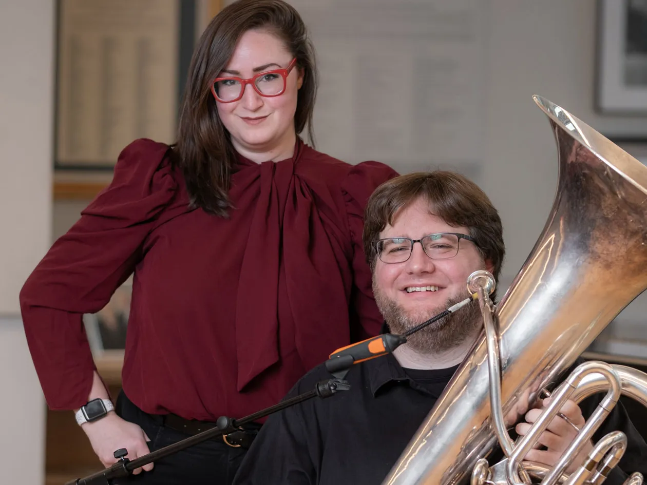 A young man smiles as he sits holding a tuba behind a microphone stand. His teacher stands close beside him with one hand on her hip. She wears bright red glasses and smiles with closed lips. She looks knowing and approving. They’re inside a music practice room.