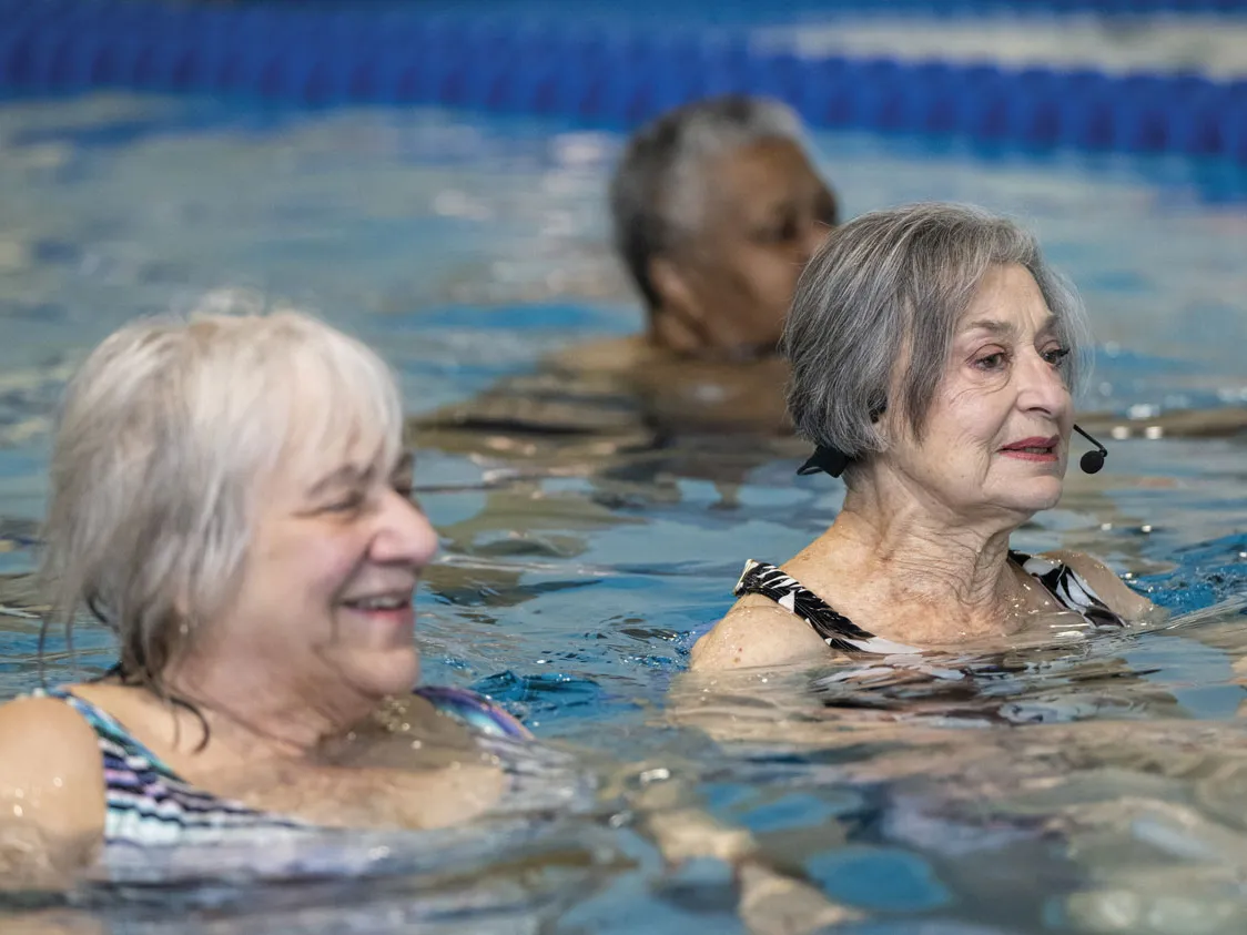 Standing neck-deep in a pool, Flo wears a headset as she calls instructions to her class. Two other people can be seen in the water with her.