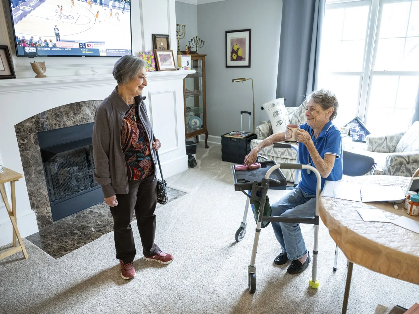 Out of her swimsuit and dressed in casual clothes, Flo stands and looks down at and old friend sitting down. A walker is in front of her friend and she holds a coffee cup. Behind them, a large wall-mounted TV plays a women's basketball game, and drapes pulled open around a large window and upholstered chairs give the room a well-decorated look.