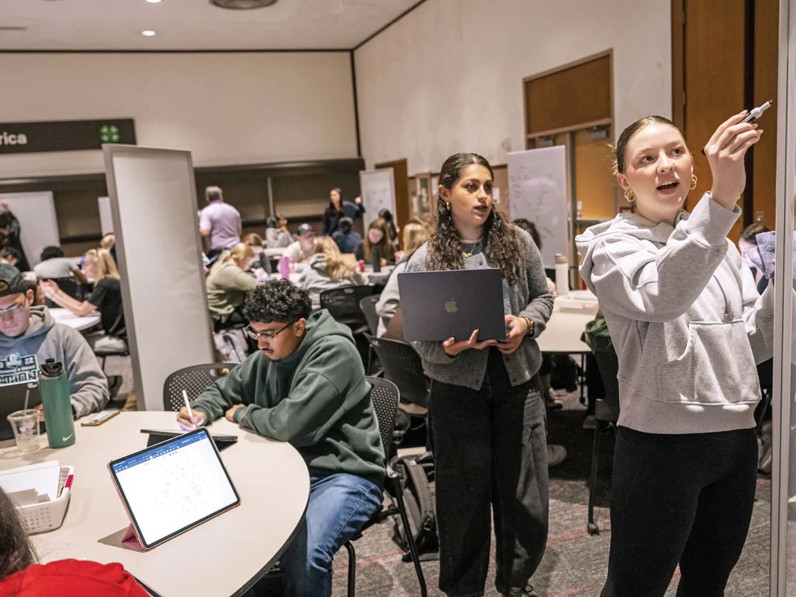 A student writes on a white board, while one more stands behind her, watching. Her formulate. At the table they stand in front of, students variously watch or check facts on their laptops. Everyone looks focused and hard at work.