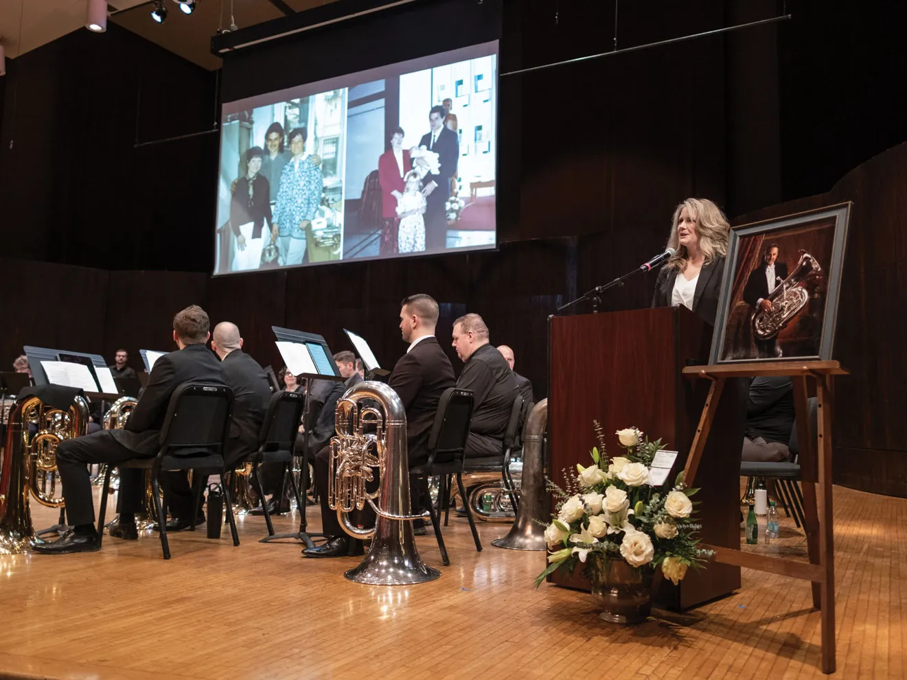 At a celebration of Jim Akins’ life, a group of musicians sit on a stage holding their instruments. To the right, his daughter speaks at a podium about her dad. His framed portrait sits on an easel nearby. Above the stage, a screen shows family photos.