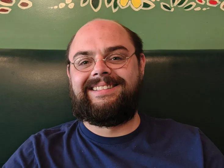 Eric Bretschneider smiles as he sits in a booth wearing a t-shirt and eyeglasses. He has dark hair, a beard and seems to be enjoying himself.