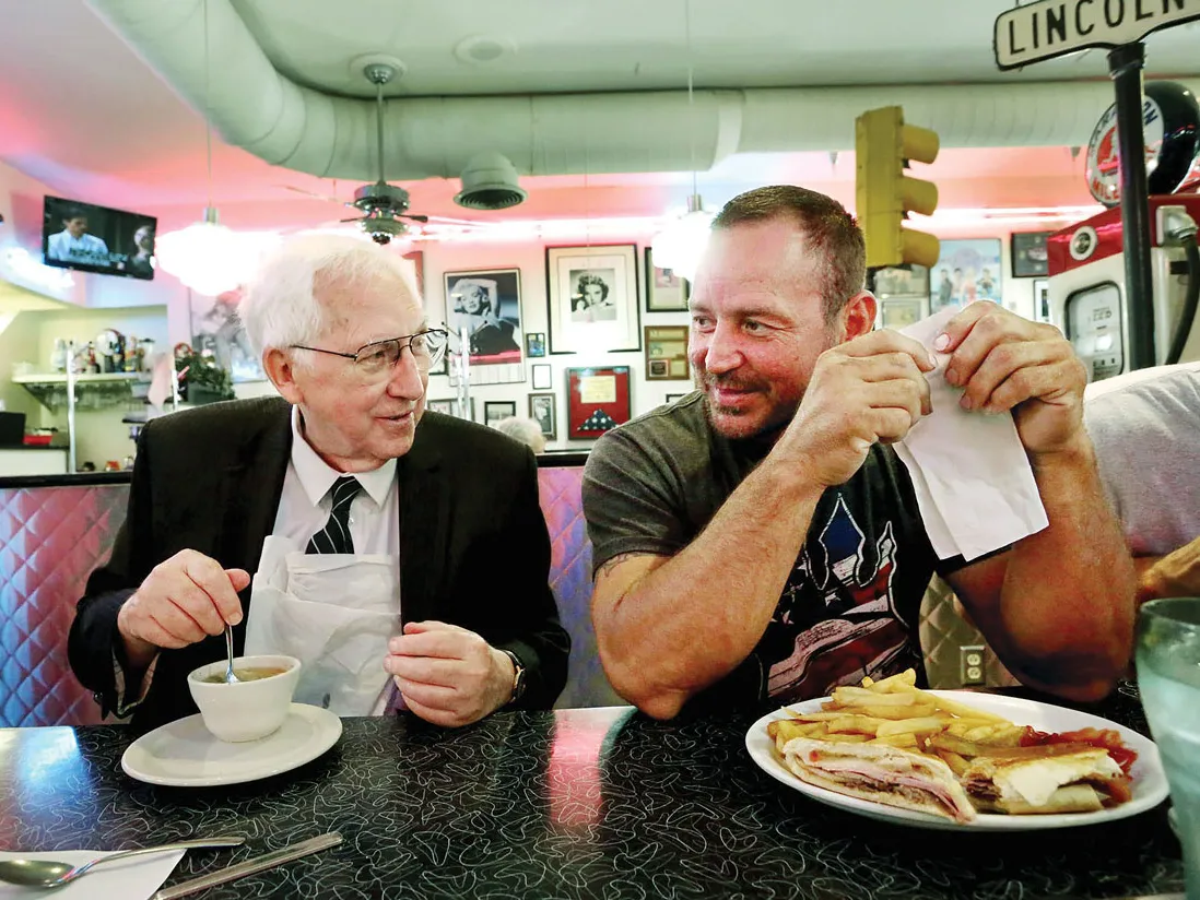 This image shows two people seated next to each other at a diner-style booth, engaged in conversation while eating. Hugh Dorrian is an older white man with white hair and a friendly, grandfatherly smile. He has a small bowl of soup. The other man is younger and wears a T-shirt and has a sandwich and fries.