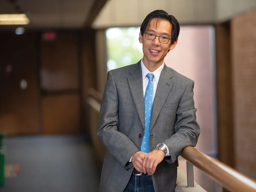 A man of Asian descent smiles as poses for this photo wearing a suit. He casually leans on a railing inside a university building. He looks like the kind of professor you'd want to take a class from--good natured and intelligent.