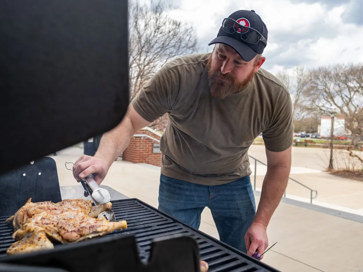 Michael Cressman leans over a grill while cooking a chicken. Cressman is a white man wearing a T-shirt, jeans and ballcap. He looks focused as he uses tongs to check the underside on Ohio State’s campus.