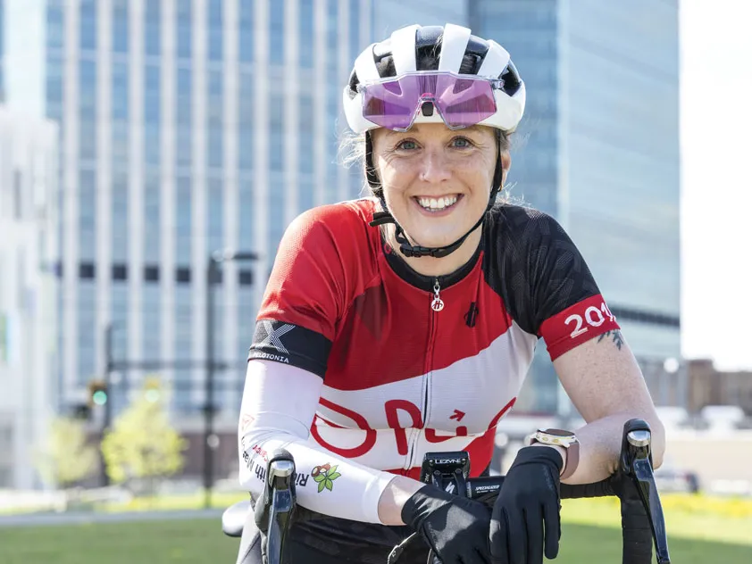 A white woman on her bicycle and wearing a riding helmet and jersey smiles as she poses for a photo in front of Ohio State's new hospital tower