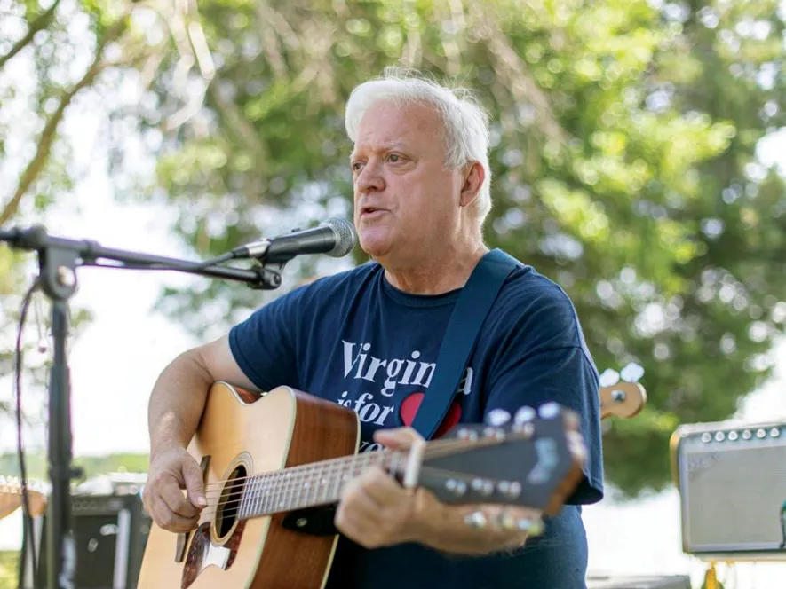 Dr. Richard Prass plays an acoustic guitar and singing into a microphone. Trees and music equipment are in the background. He’s an older white man with white hair and T-shirt that probably says “Virginia is for lovers”