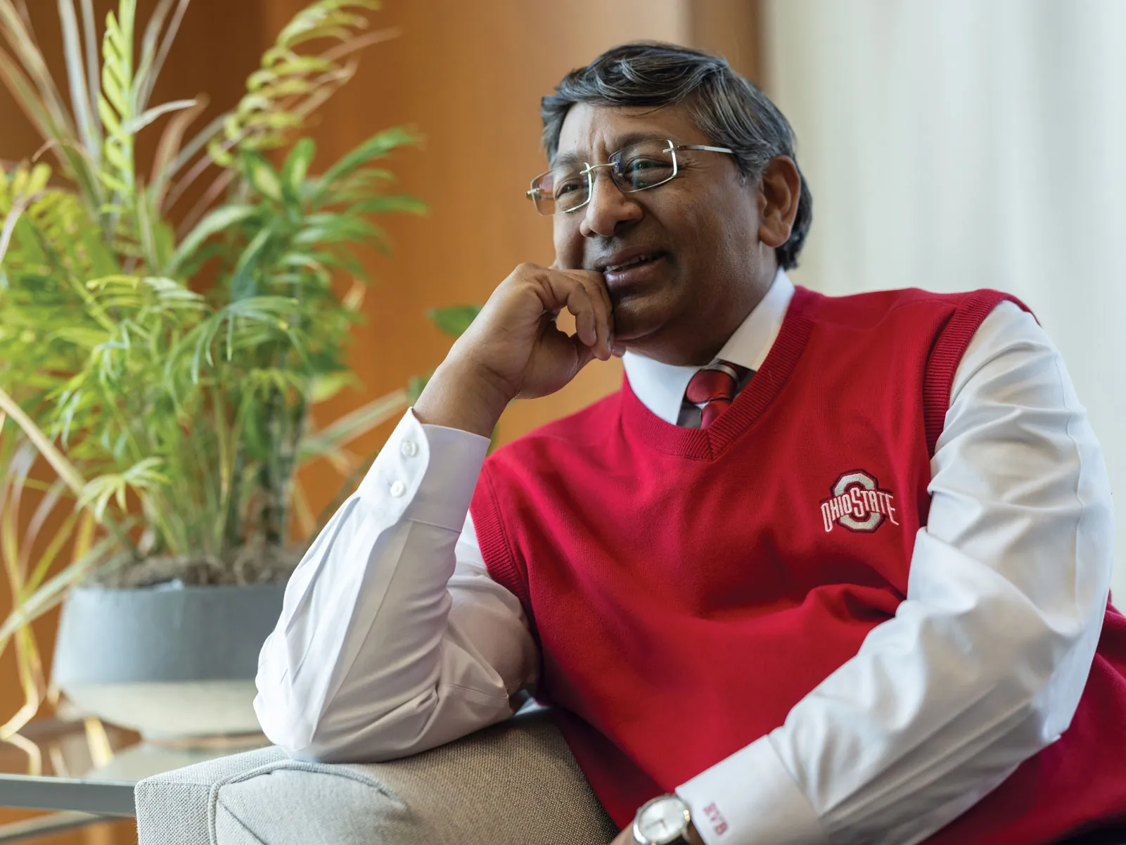 Ravi Bellamkonda leans one elbow on the arm of a couch as he leans into it and chuckles. He wears a scarlet Ohio State sweater vest, glasses and has salt-and-pepper tussled hair. He appears to be invested in a conversation
